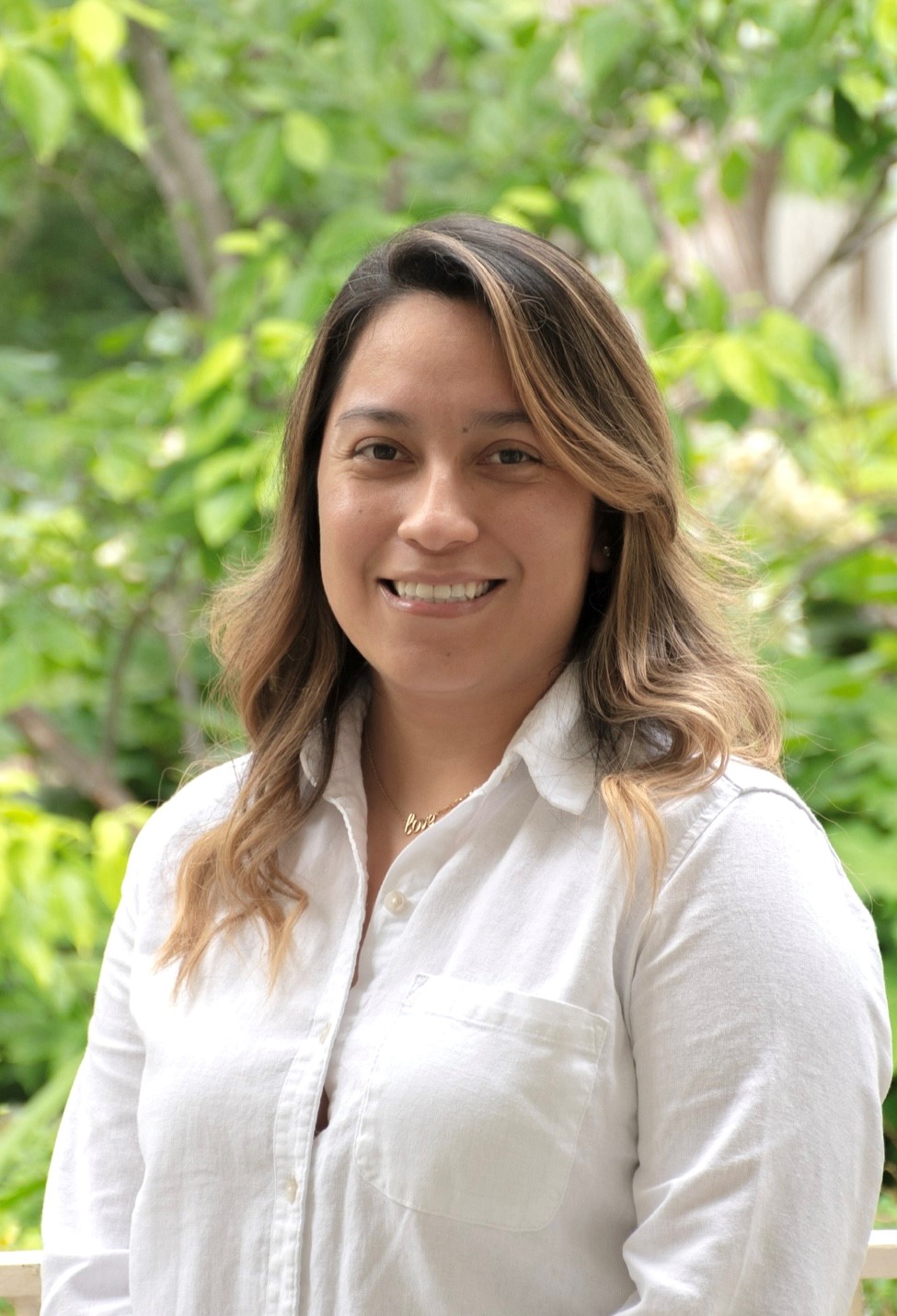 Smiling woman with curly hair outdoors, wearing a white shirt.